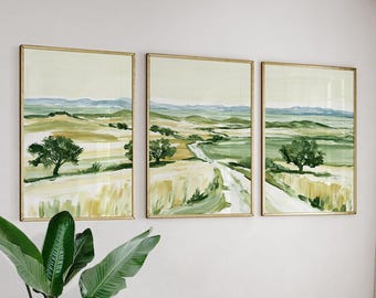May include: A triptych of three framed landscape paintings featuring a rolling countryside with green fields, golden wheat, scattered trees, and a winding dirt path under a pale sky, all enclosed in thin gold-toned metal frames.