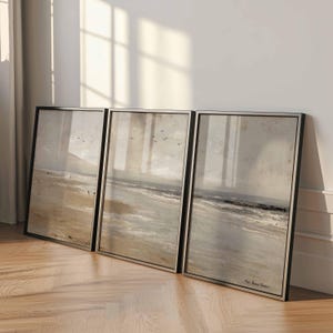May include: Three framed landscape art prints depicting a beach scene with a muted color palette of beige, gray, and black. The artwork features a shoreline, ocean waves, and a cloudy sky. The frames are black with silver borders.