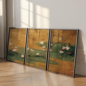 May include: Three framed art prints featuring a traditional Japanese painting style. The artwork depicts white cherry blossoms and green water features against a gold background. The frames are black with silver trim.