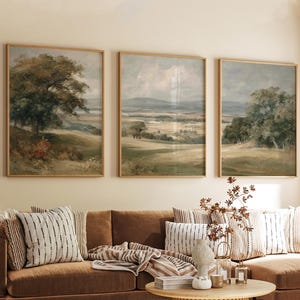 Puede incluir: Tres pinturas de paisajes enmarcadas de estilo vintage cuelgan sobre un sofá marrón. La obra de arte representa colinas, árboles y un cielo nublado en tonos apagados de verde, marrón y gris. Los marcos son de color madera clara.