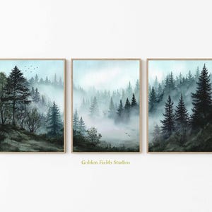 Könnte beinhalten: Drei gerahmte Aquarellmalereien einer nebligen Waldszene. Das Kunstwerk zeigt hohe immergrüne Bäume in Grün- und Blautönen mit weichem, atmosphärischem Nebel. Die Rahmen sind hellbraun. Der Text "Golden Fields Studios" ist sichtbar.