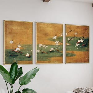 May include: Set of three framed art prints featuring a traditional Japanese painting style. Each print depicts a scene with white cherry blossoms, green water, and a golden background. The frames have a gold finish.