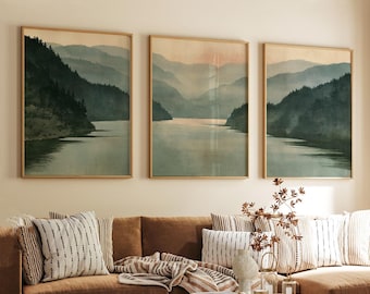 Op de afbeelding: Een set van drie rechthoekige wandkunstprints in licht houten lijsten, elk met een sereen landschap van donkergroene bergen en bossen die weerspiegelen in een kalm wateroppervlak onder een zachte, wazige lucht in gedempte beige en saliegroene tinten.