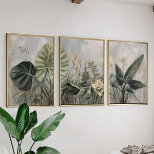 Könnte beinhalten: Dreiteiliges Set gerahmter botanischer Kunstdrucke. Jeder Druck zeigt eine andere Anordnung von grünen und gelben Blättern vor einem grauen Hintergrund. Die Rahmen sind goldfarben und verleihen dem Kunstwerk Eleganz.