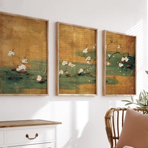 May include: Three framed wall art pieces. Each features a vintage-style painting of white cherry blossoms over a green and gold water scene. The frames are made of light-colored wood, and the art is displayed on a white wall.