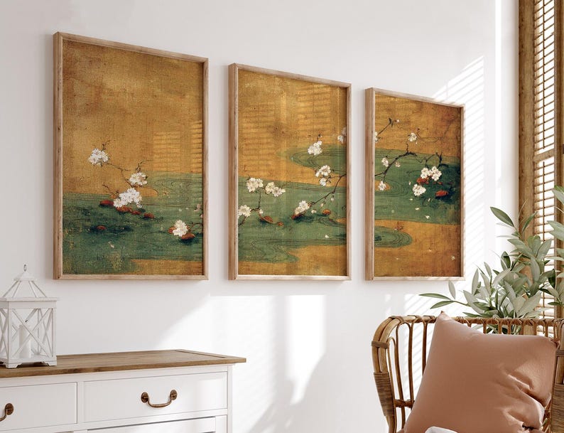 May include: Three framed wall art pieces. Each features a vintage-style painting of white cherry blossoms over a green and gold water scene. The frames are made of light-colored wood, and the art is displayed on a white wall.
