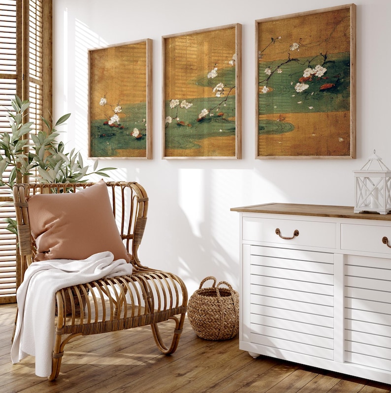 May include: Three framed wall art pieces with a Japanese-inspired design. Each piece features a gold background with a green and white floral motif. The art is displayed above a white cabinet and a wicker chair.