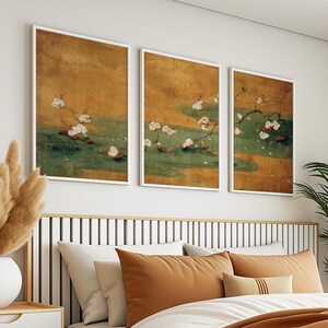 May include: Three framed art prints featuring a traditional Japanese painting style. Each print displays a branch of white blossoms over a green and gold background. The artwork is displayed above a bed with orange and white pillows.