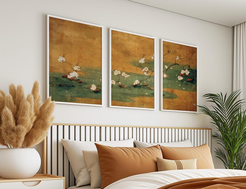 May include: Three framed art prints featuring a traditional Japanese painting style. Each print displays a branch of white blossoms over a green and gold background. The artwork is displayed above a bed with orange and white pillows.