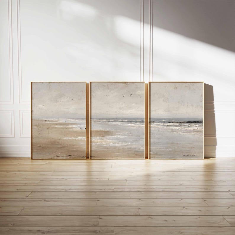 May include: Three framed beach landscape paintings in neutral tones. The artwork depicts a sandy beach, ocean waves, and a cloudy sky. The frames are a light gold color, and the paintings are displayed on a wooden floor.
