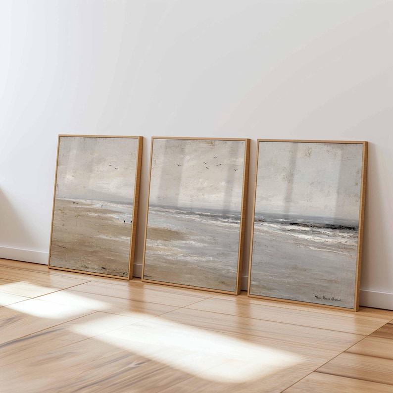 May include: Three framed beach landscape paintings. Each artwork features a muted color palette of beige, gray, and blue, depicting a shoreline with subtle brushstrokes. The frames are a light wood tone, adding a natural touch to the coastal theme.