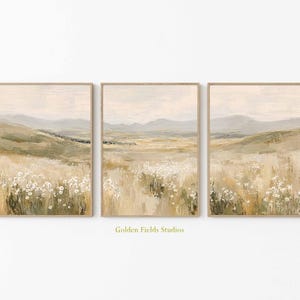 Op de afbeelding: Set van drie ingelijste landschapskunstprints. Elke print toont een veld met wilde bloemen op de voorgrond, glooiende heuvels en een gedempte lucht. Het kunstwerk heeft een neutraal kleurenpalet met beige, groene en grijze tinten.
