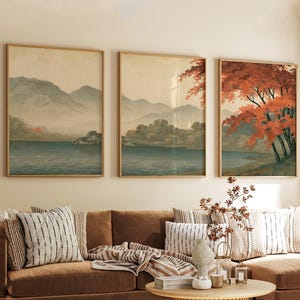 Könnte beinhalten: Ein Set aus drei gerahmten Wandbildern, die eine durchgehende Landschaftsszene zeigen. Das Kunstwerk stellt einen ruhigen See, ferne bläuliche Berge und Bäume mit leuchtend orangefarbenem und rotem Herbstlaub in hellen Holzrahmen dar.