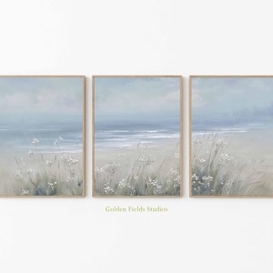 Puede incluir: Tres impresiones artísticas enmarcadas que representan una serena escena de playa. La obra de arte presenta una paleta de colores suaves de azules, grises y beige, con un enfoque en el océano, el cielo y las hierbas costeras. Los marcos son de color madera clara. El texto "Golden Fields Studios" es visible.