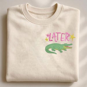 May include: Cream-colored sweatshirt with the word "LATER" in pink script, accented by yellow stars. A green alligator graphic is below the text. The sweatshirt is folded neatly.