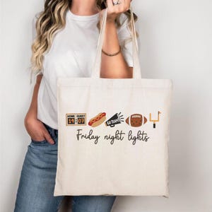 Op de afbeelding: Een cr&egrave;mekleurige canvas draagtas met een grafische print van een scorebord met 14:07, een hotdog, een megafoon met de tekst GO TEAM!, een football en een doelpaal. Onder de iconen staat de tekst Friday night lights in zwarte letters gedrukt.