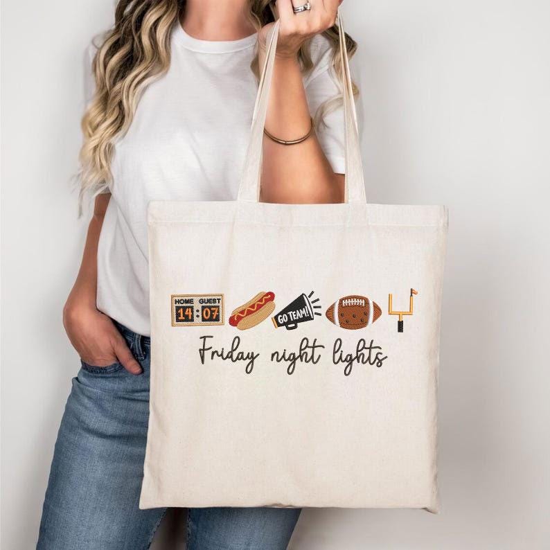 Op de afbeelding: Een cr&egrave;mekleurige canvas draagtas met een grafische print van een scorebord met 14:07, een hotdog, een megafoon met de tekst GO TEAM!, een football en een doelpaal. Onder de iconen staat de tekst Friday night lights in zwarte letters gedrukt.