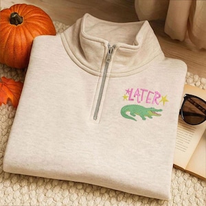 May include: A light beige sweatshirt with a quarter-zip neckline. The sweatshirt features the word "LATER" in pink with yellow star accents, and a green alligator graphic. A pumpkin and sunglasses are in the background.