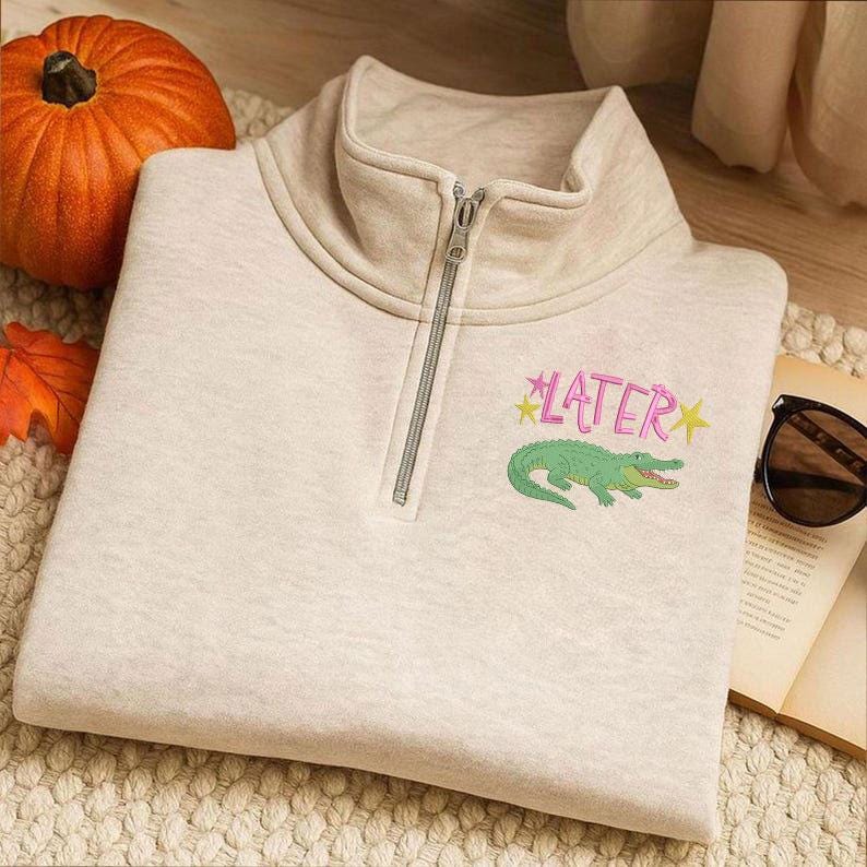 May include: A light beige sweatshirt with a quarter-zip neckline. The sweatshirt features the word "LATER" in pink with yellow star accents, and a green alligator graphic. A pumpkin and sunglasses are in the background.