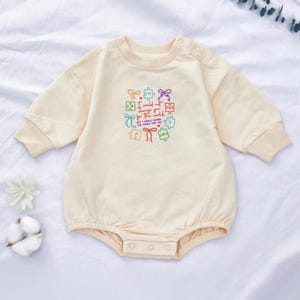 May include: Cream-colored baby romper with long sleeves and a round neckline. The romper features a colorful embroidered design of puzzle pieces and the text "I would not change you for the world, but I would change the world for you."