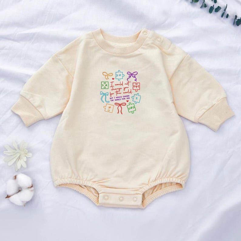 May include: Cream-colored baby romper with long sleeves and a round neckline. The romper features a colorful embroidered design of puzzle pieces and the text "I would not change you for the world, but I would change the world for you."