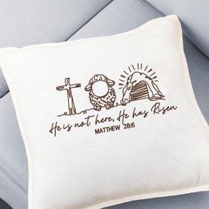 K&ouml;nnte beinhalten: Cremefarbenes quadratisches Kissen mit einer braunen Strichzeichnung eines Kreuzes, eines Schafs und eines leeren Grabes mit dem Text "He is not here, He has Risen" und "MATTHEW 28:6". Das Kissen liegt auf einer hellblauen Oberfl&auml;che.