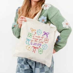 May include: Cream-colored tote bag with a colorful design featuring puzzle pieces and bows. The text on the bag reads, "I would not change you for the world, but I would change the world for you." The bag has a simple, minimalist design.