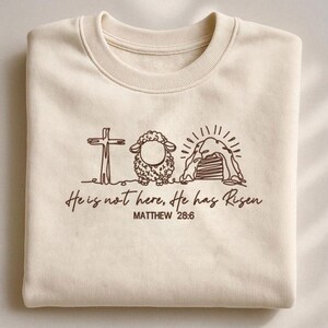 K&ouml;nnte beinhalten: Cremefarbenes Sweatshirt mit brauner Grafik eines Kreuzes, Schafs und Grabes. Der Text "He is not here, He has Risen" und "MATTHEW 28:6" sind darunter gedruckt. Das Sweatshirt ist gefaltet.