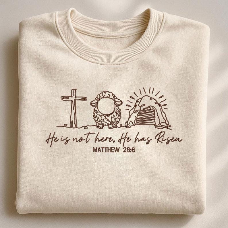 K&ouml;nnte beinhalten: Cremefarbenes Sweatshirt mit brauner Grafik eines Kreuzes, Schafs und Grabes. Der Text "He is not here, He has Risen" und "MATTHEW 28:6" sind darunter gedruckt. Das Sweatshirt ist gefaltet.