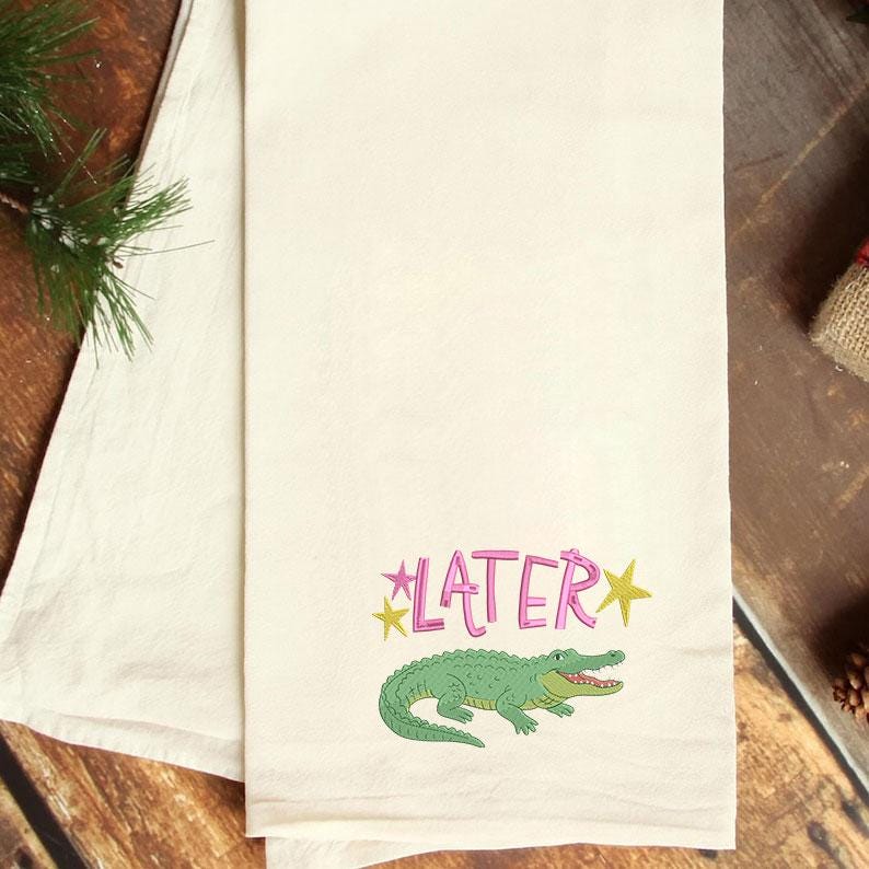 May include: Off-white kitchen towel with a green alligator illustration and the word "LATER" in pink with yellow star accents. The towel is folded and has a simple, clean design.