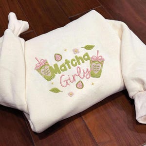 May include: Cream-colored sweatshirt with embroidered "Matcha Girly" design. Features two matcha drink illustrations, leaves, flowers, and a strawberry. The design is in shades of green, pink, and yellow.