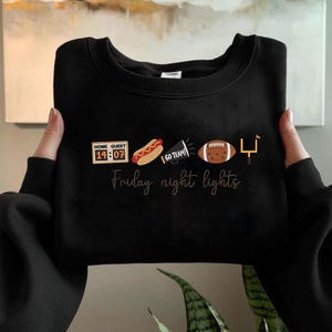 Op de afbeelding: Een zwart sweatshirt met ronde hals en een grafische print met een scorebord dat 14:07 aangeeft, een hotdog, een megafoon met de tekst GO TEAM!, een football en een doelpaal. Onder de iconen staat de tekst Friday night lights in een cursief lettertype.