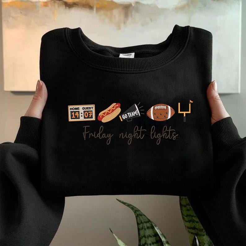 Op de afbeelding: Een zwart sweatshirt met ronde hals en een grafische print met een scorebord dat 14:07 aangeeft, een hotdog, een megafoon met de tekst GO TEAM!, een football en een doelpaal. Onder de iconen staat de tekst Friday night lights in een cursief lettertype.