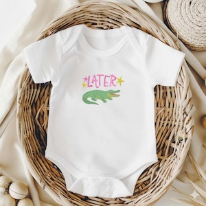 May include: White baby bodysuit with a green alligator graphic and the word "LATER" in pink with yellow star accents. The bodysuit is short-sleeved and made of soft material. The bodysuit is displayed in a woven basket.