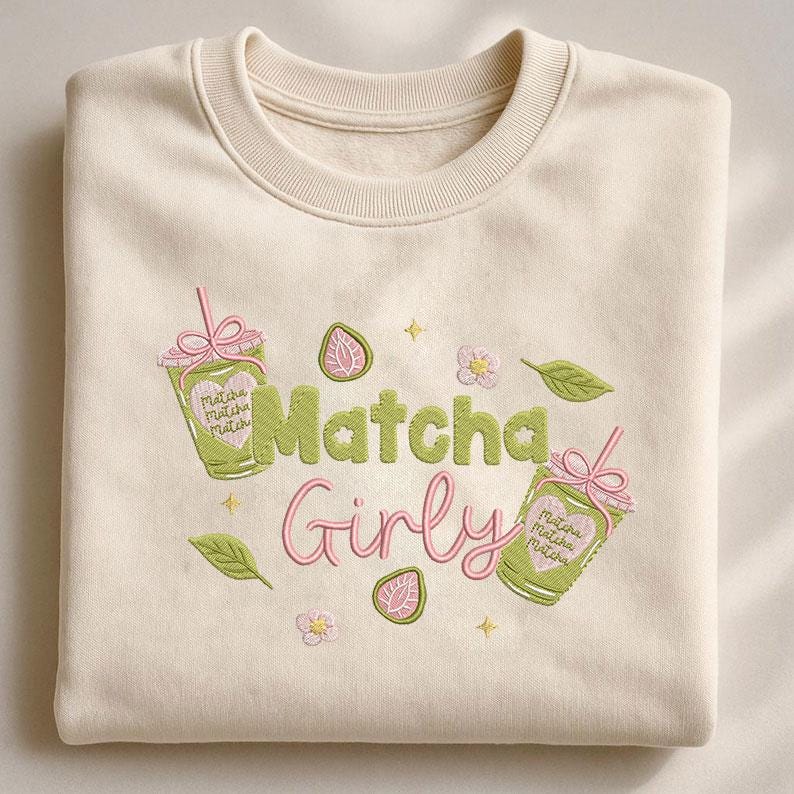 May include: Cream-colored sweatshirt with embroidered design. The design features the words "Matcha Girly" in green and pink, along with matcha drink illustrations, flowers, and leaves. The sweatshirt has a crew neck and long sleeves.