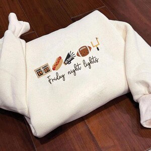 Op de afbeelding: Een cr&egrave;mekleurig sweatshirt met geborduurde iconen, waaronder een scorebord met 14:07, een hotdog, een megafoon met de tekst GO TEAM!, een football en een doelpaal, met daaronder de in script geborduurde tekst Friday night lights.