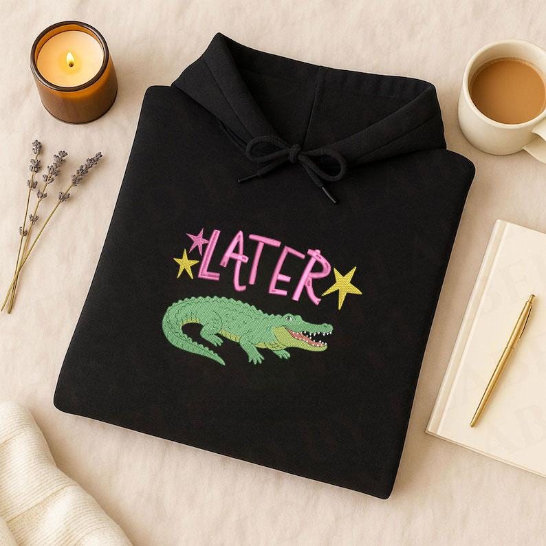 May include: Black hoodie with a drawstring hood featuring the word "LATER" in pink with yellow stars and a green alligator graphic. The hoodie is made of a soft material.