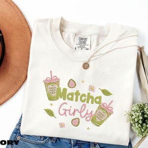 May include: Ivory-colored t-shirt with the words "Matcha Girly" in green and pink script. The shirt features matcha drink illustrations and floral accents. A gold necklace and a brown hat are also visible.