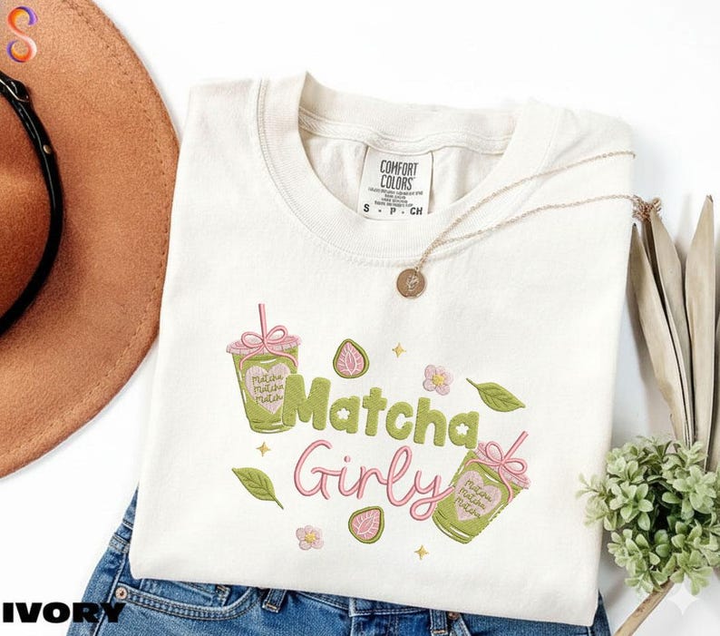 May include: Ivory-colored t-shirt with the words "Matcha Girly" in green and pink script. The shirt features matcha drink illustrations and floral accents. A gold necklace and a brown hat are also visible.