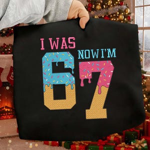 May include: Black tote bag with the text "I WAS 67 NOW I'M 67". The numbers are designed to look like donuts with blue and pink icing and sprinkles. The text "I WAS" is pink, and "NOW I'M" is blue.
