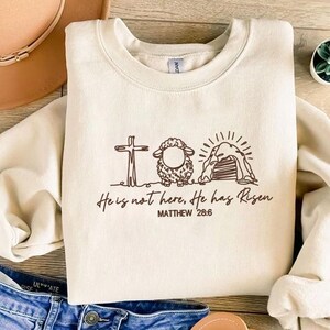 K&ouml;nnte beinhalten: Cremefarbenes Sweatshirt mit brauner Grafik eines Kreuzes, Schafs und Grabes mit dem Text "He is not here, He has Risen" und "MATTHEW 28:6". Das Sweatshirt ist aus einem weichen Material.