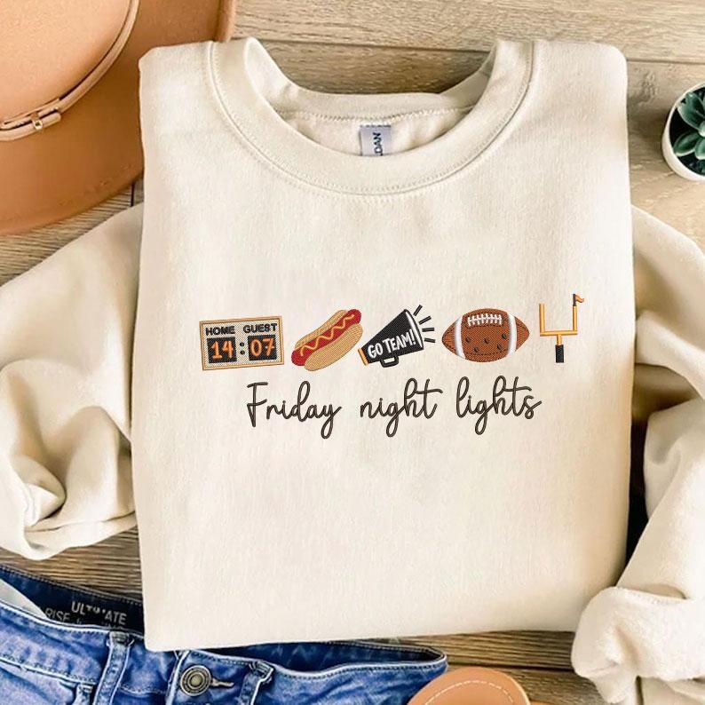 Op de afbeelding: Een cr&egrave;mekleurige sweater met ronde hals en geborduurde iconen van een scorebord met 14:07, een hotdog, een megafoon met de tekst GO TEAM!, een football en een doelpaal, met daaronder de cursieve tekst Friday night lights.