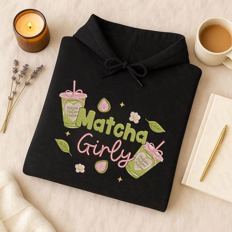 May include: Black hoodie with the embroidered text "Matcha Girly" in green and pink. The design includes matcha drinks, flowers, and leaves. A candle, a cup of coffee, and a notebook with a pen are also visible.