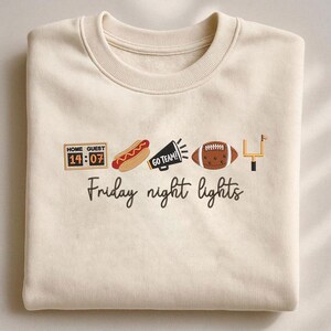 Op de afbeelding: Een cr&egrave;mekleurige sweater met ronde hals en geborduurde iconen, waaronder een scorebord met 14:07, een hotdog, een megafoon met de tekst GO TEAM!, een football en een doelpaal, met de tekst Friday night lights eronder geborduurd.