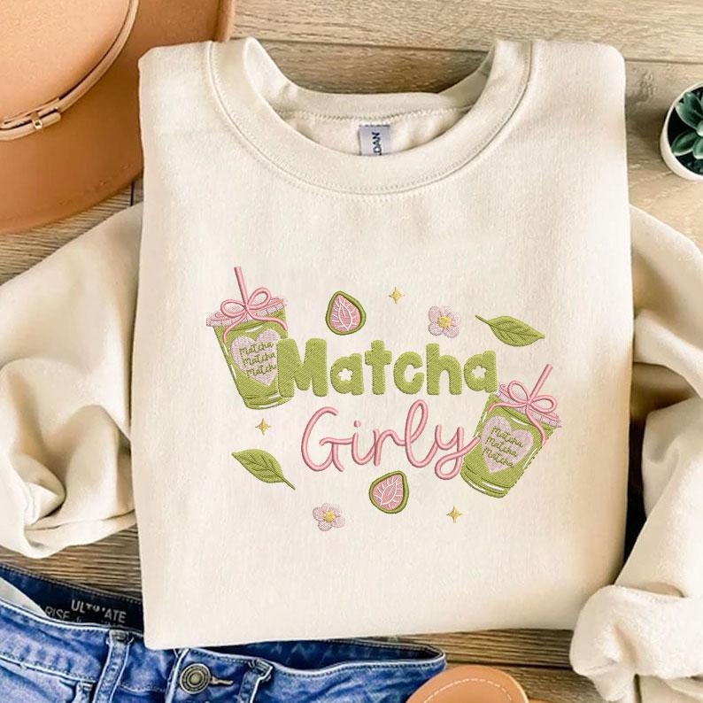 May include: Cream-colored sweatshirt with the text "Matcha Girly" in green and pink. The design includes matcha drinks, strawberries, flowers, and leaves. The sweatshirt is folded on a wooden surface.
