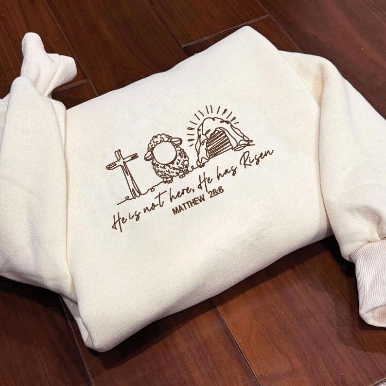 K&ouml;nnte beinhalten: Cremefarbenes Sweatshirt mit brauner Grafik, die ein Kreuz, ein Schaf und ein offenes Grab zeigt. Der Text "He is not here, He has Risen" und "MATTHEW 28:6" sind darunter aufgedruckt. Das Sweatshirt hat gerippte B&uuml;ndchen und einen Rundhalsausschnitt.