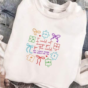 May include: White sweatshirt with colorful puzzle pieces and bows. The text reads, "I would not change you for the world, but I would change the world for you."