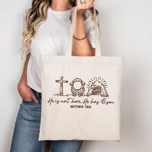 K&ouml;nnte beinhalten: Beige Tragetasche mit einer braunen Strichzeichnung eines Kreuzes, Schafs und Grabes. Der Text lautet "He is not here, He has Risen" und "Matthew 28:6". Die Tasche hat zwei Griffe und wird von einer Person gehalten.