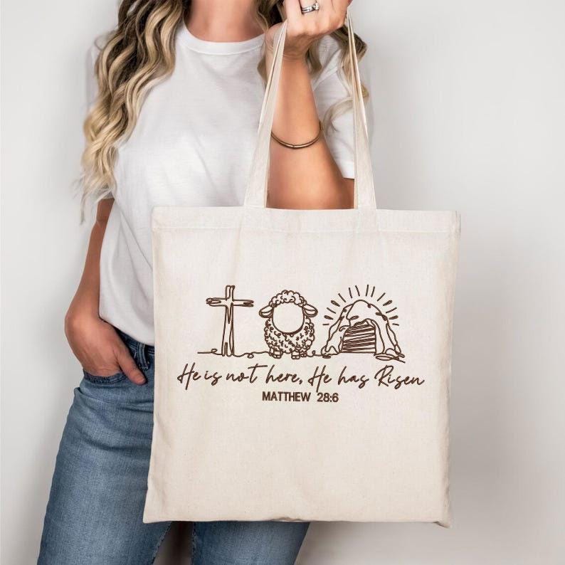 K&ouml;nnte beinhalten: Beige Tragetasche mit einer braunen Strichzeichnung eines Kreuzes, Schafs und Grabes. Der Text lautet "He is not here, He has Risen" und "Matthew 28:6". Die Tasche hat zwei Griffe und wird von einer Person gehalten.