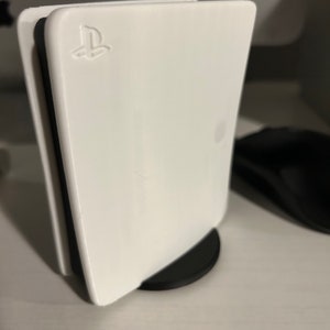 May include: A white PlayStation 5 console with the PlayStation logo on the front. The console is on a black stand.